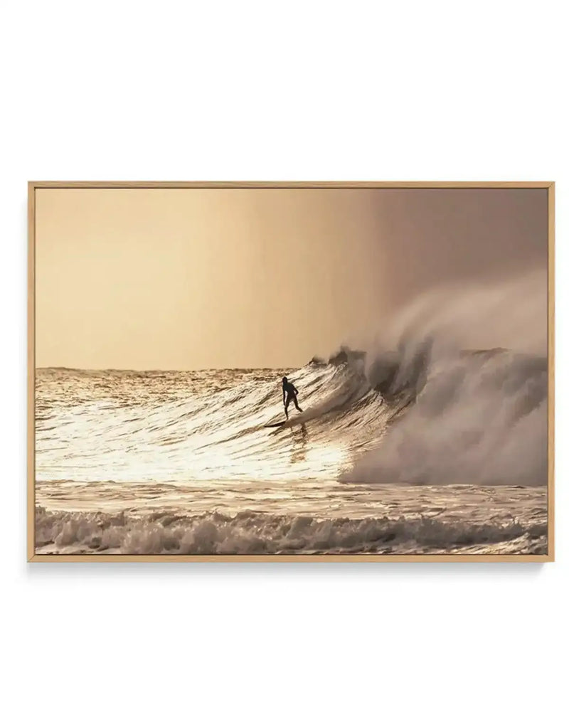 Framed canvas art print featuring a sepia-toned photograph of a surfer in a wetsuit riding a large wave at Bells Beach during golden hour. The image captures the surfer in profile against a towering wave with spray and mist, with smaller whitecaps visible in the foreground. The warm, muted palette transitions from pale cream sky to soft brown and tan tones across the water and sand. The photograph is mounted in a natural wood frame with a visible border.