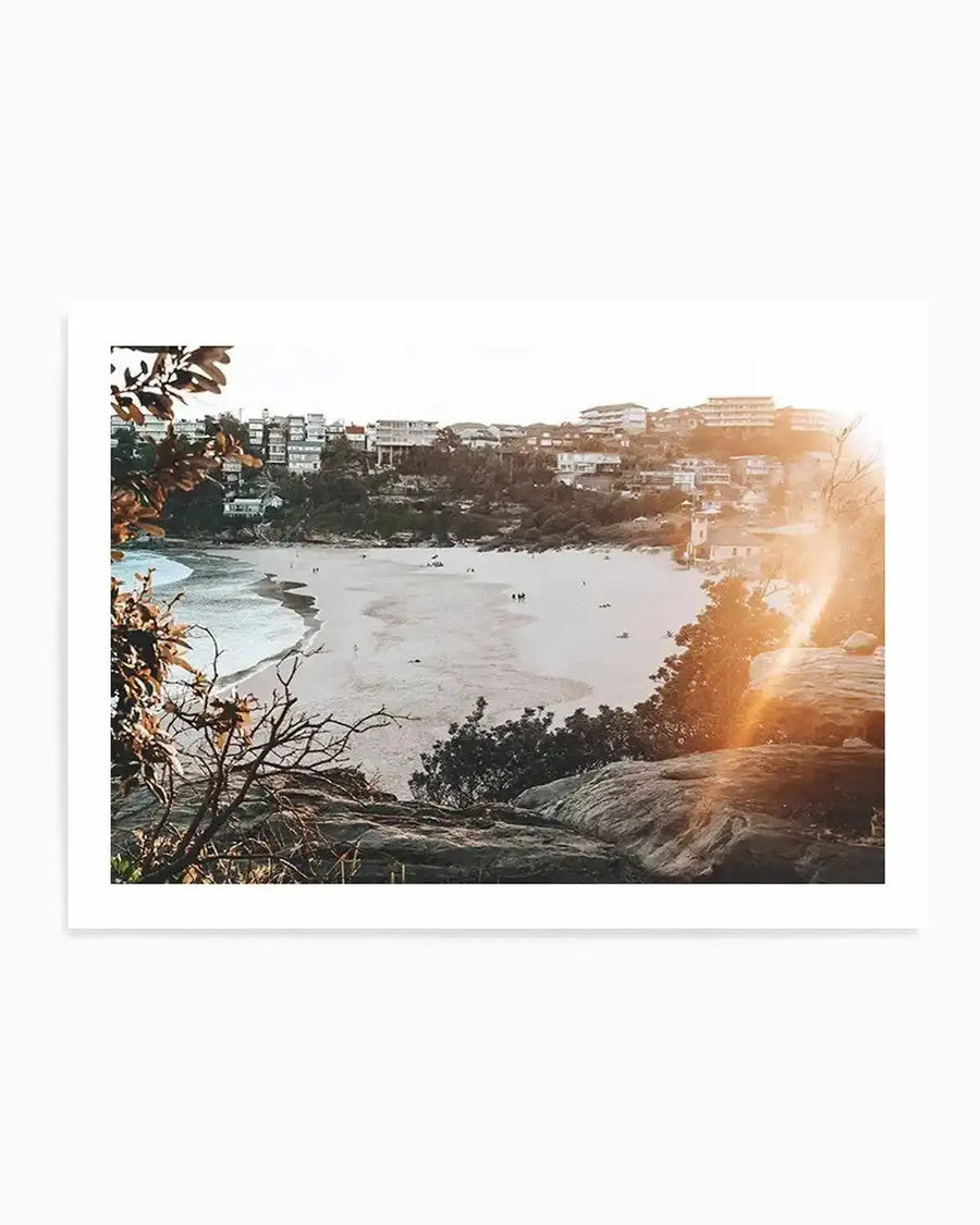 Framed photography art print capturing a sun-drenched coastal beach scene from an elevated perspective, with a natural wood frame and white border. The composition features a crescent-shaped sandy beach with gentle waves, surrounded by lush green hills dotted with houses, all bathed in the warm glow of a bright sun flare on the right.