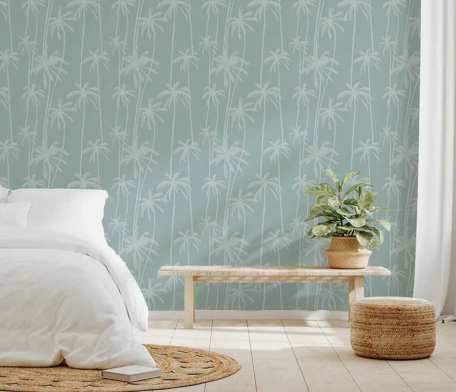 Lifestyle shot of a bedroom featuring "Good Palms Bondi" peel and stick wallpaper. The wallpaper has a repeating pattern of hand-drawn palm trees in a light, almost white, grey against a soft, muted blue-green background. The room includes a bed with white linen bedding, a light wood slatted bench, a woven jute rug, a potted plant in a woven basket, and a woven jute ottoman.