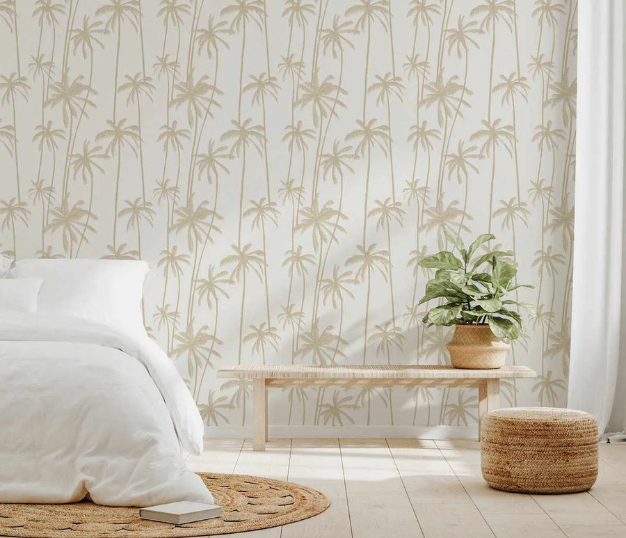"Good Palms Bronzed wallpaper featuring a repeating pattern of tall, slender palm trees with full fronds in a warm, muted bronzed beige against a soft white background, displayed on a bedroom wall. The room includes a white bed with white bedding, a light wood slatted bench with a woven top, a potted green plant in a woven basket, a round woven rug, and a woven cylindrical ottoman."