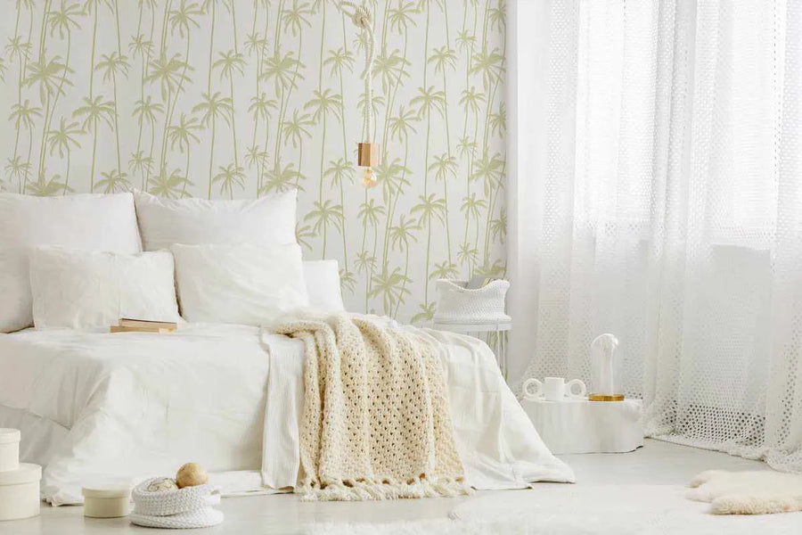 Lifestyle shot of a bright bedroom featuring Good Palms Pistachio Wallpaper. The wallpaper has a repeating pattern of hand-drawn palm trees in a soft pistachio green on a crisp white background, creating a fresh, tropical feel. The wallpaper is visible behind a bed dressed in all white linen with a cream knitted throw blanket. A light fixture with a wooden accent hangs from the ceiling, and sheer white curtains adorn the window on the right.