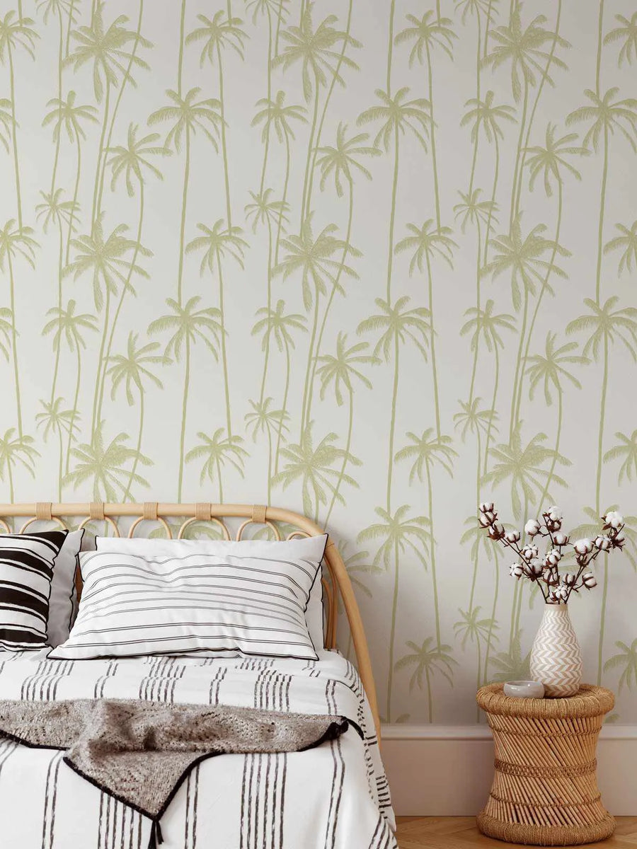 "Good Palms Pistachio Wallpaper featuring a repeating pattern of hand-drawn palm trees in a soft pistachio green on a white background, creating a tropical and serene atmosphere in a bedroom. The wallpaper is visible behind a rattan bed frame with white and black striped bedding. To the right, a small rattan side table holds a vase with cotton branches."
