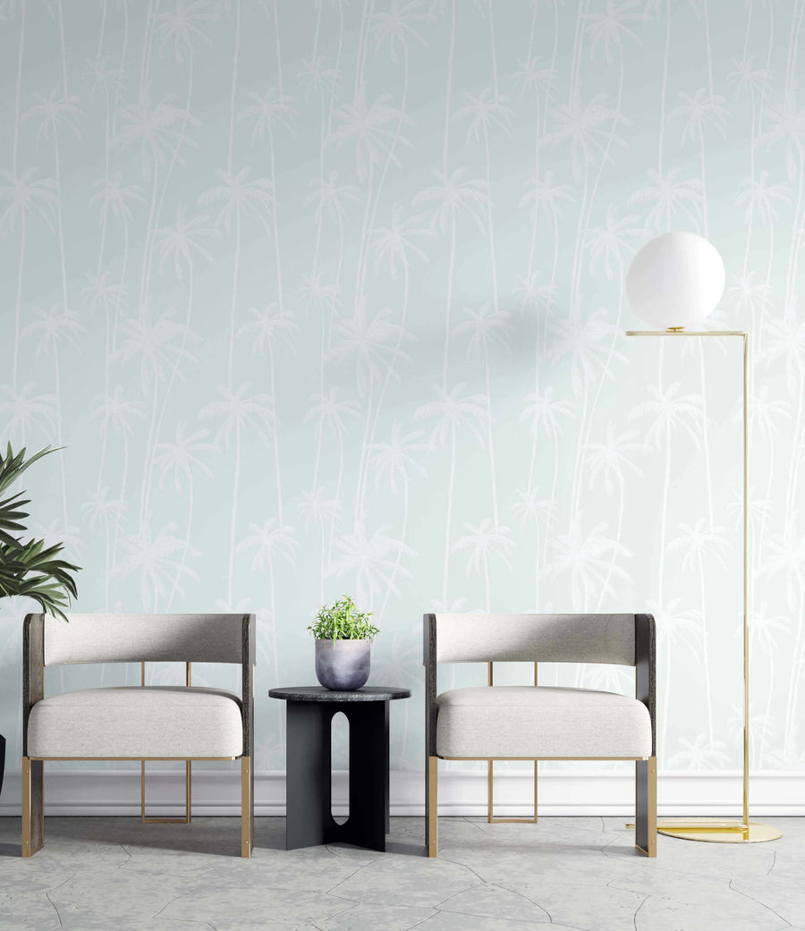 Lifestyle shot of a modern living space featuring Good Palms Sage Wallpaper. The wallpaper has a repeating pattern of white palm trees with thin trunks and full fronds on a soft sage green background, creating a subtle tropical motif. The room includes two light grey upholstered chairs with dark wood and gold metal frames, a small black side table with a potted green plant, and a tall gold floor lamp with a spherical white shade. The floor is light grey with a subtle textured pattern.