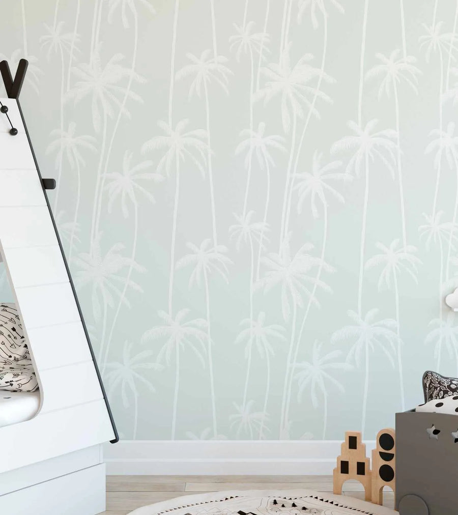 Lifestyle shot of a child's bedroom featuring wallpaper with a repeating pattern of tall, slender palm trees in white against a soft sage green background. The wallpaper covers the entire wall, creating a tropical yet minimalist feel. In the foreground, a white A-frame bed is partially visible on the left, and a grey toy box with star cutouts and wooden building blocks are on the right. A round rug with a black and white geometric pattern is on the light wood floor.