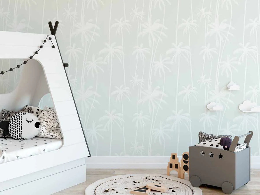 Lifestyle shot of a child's bedroom featuring Good Palms Sage wallpaper. The wallpaper has a repeating pattern of white palm trees with thin trunks and full fronds on a soft sage green background. The room includes a white teepee-style bed with black accents and string lights, a grey wheeled toy bin with wooden blocks, and a circular white rug with a black pattern. White cloud-shaped shelves are mounted on the wall.