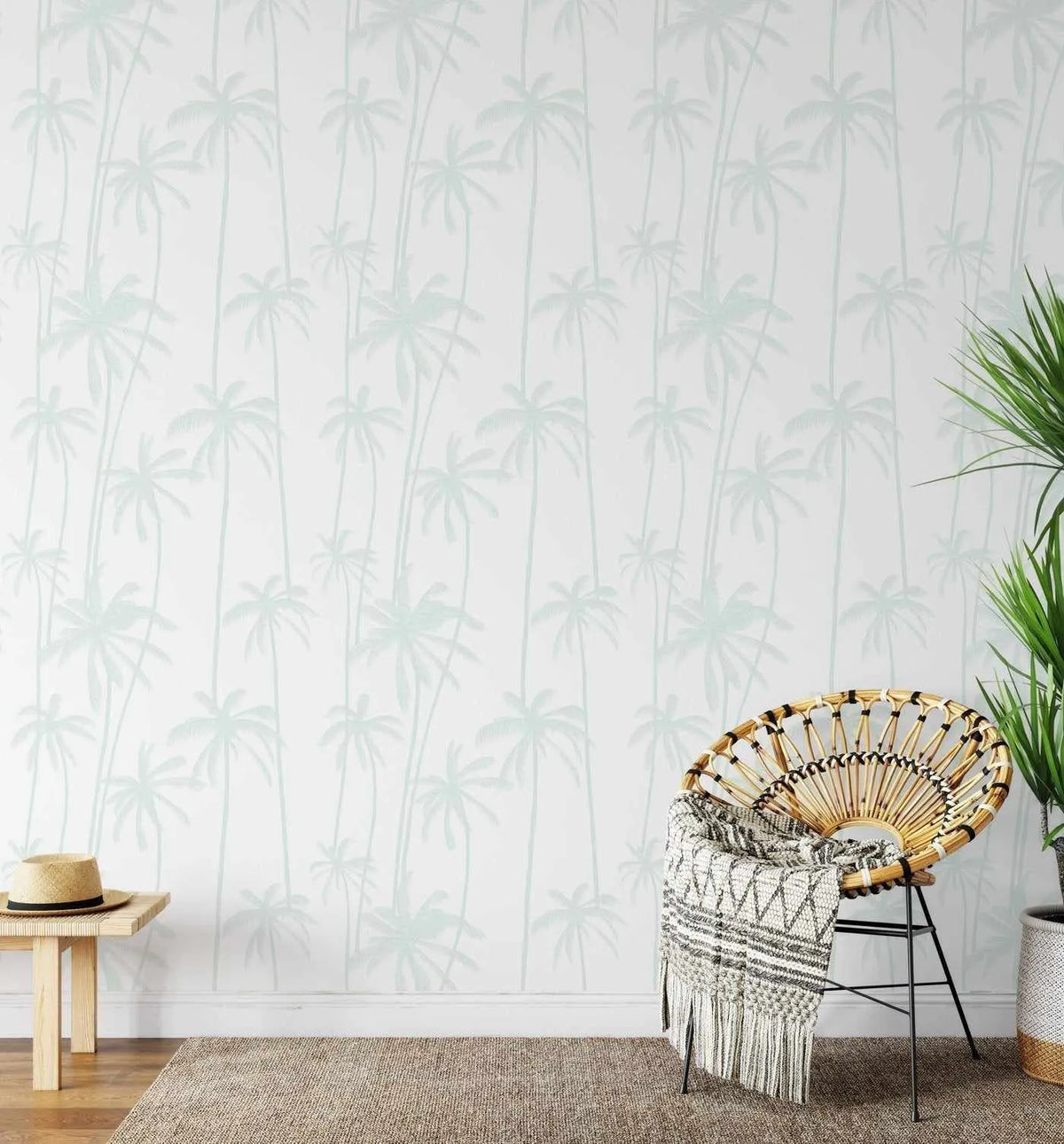 Good Palms Seafoam Wallpaper displayed on a wall in a bright room. The wallpaper features a repeating pattern of tall palm trees in soft seafoam green and pale grey-green tones on a white background with subtle vertical striping. The illustration style is delicate and botanical. In the foreground, a woven rattan chair with a cream macramé throw, a small wooden side table with a straw hat, a jute area rug, potted plants in woven baskets, and green foliage complete the serene, coastal-inspired setting.