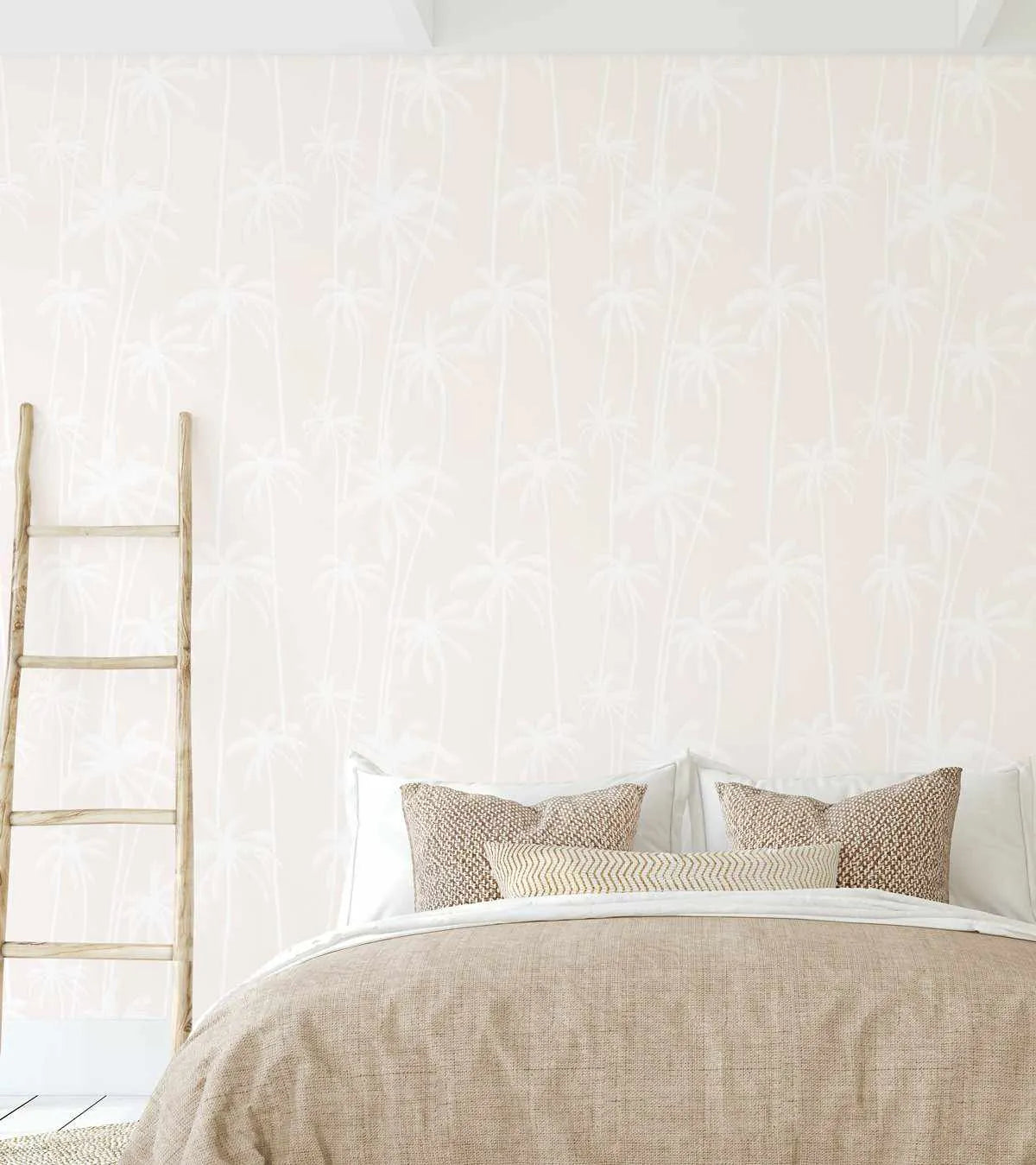 "Good Palms Soft Sand Wallpaper with a repeating pattern of hand-drawn palm trees in a soft white against a pale, sandy beige background. The wallpaper is displayed on a bedroom wall above a bed dressed in neutral-toned bedding and pillows, with a rustic wooden ladder leaning against the wall on the left."