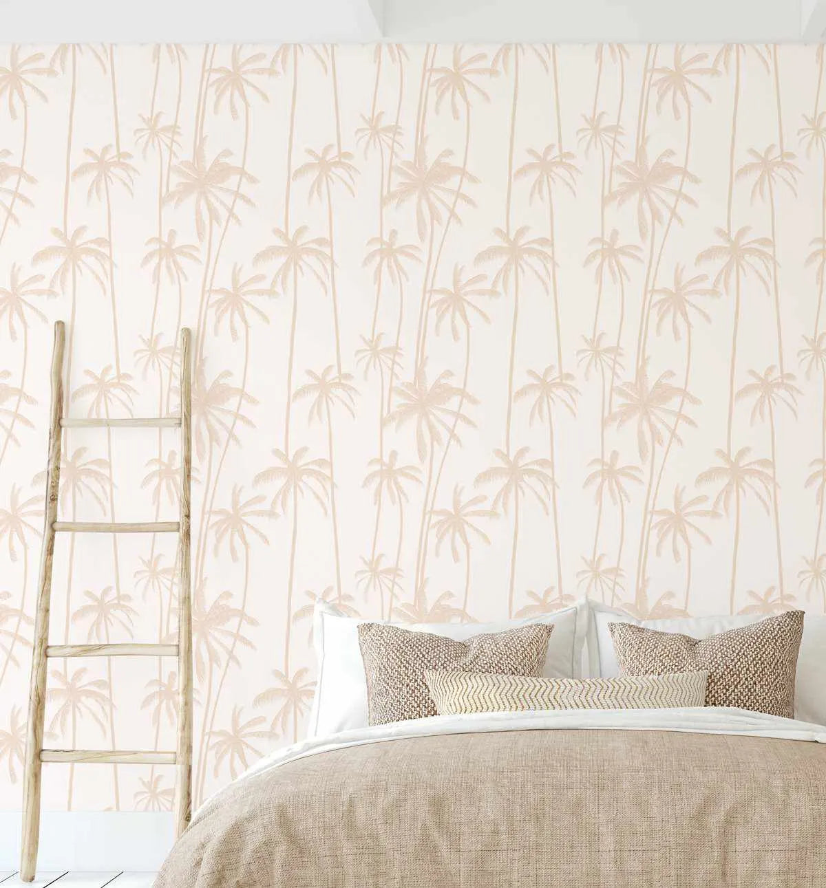 Bedroom wall featuring Good Palms Soft Terracotta Wallpaper, displaying a repeating pattern of tall palm trees in soft terracotta and beige tones on a cream background. The design shows delicate, hand-drawn style palm silhouettes arranged in vertical columns. A wooden ladder leans against the wall on the left, and a bed with neutral linen bedding and patterned throw pillows is positioned in front of the wallpapered wall, demonstrating the wallpaper's full-room application and warm, tropical aesthetic.