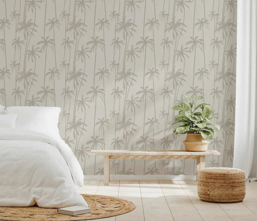 Lifestyle shot of a bedroom featuring Good Palms True Neutral Wallpaper. The wallpaper has a repeating pattern of hand-drawn palm trees in a soft, muted beige-grey against a light cream background, creating a subtle tropical motif. The room includes a white bed with white linen bedding, a light wooden bench with a woven seat, a potted plant in a woven basket, a round jute rug, and a woven jute ottoman.