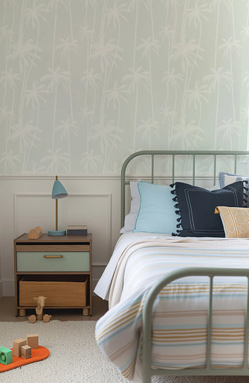 Lifestyle shot of a child's bedroom featuring a wallpaper with a repeating pattern of hand-drawn palm trees in white on a soft sage green background. The wallpaper covers the upper half of the wall above white wainscoting. A sage green metal bed frame with a light blue pillow, a dark navy pillow with tassels, and a striped duvet in white, light blue, and mustard yellow is visible. A wooden nightstand with a sage green drawer and a woven basket is next to the bed, holding a light blue lamp.