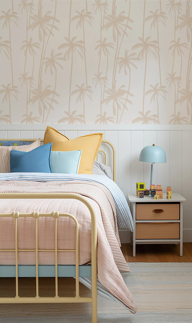 Good Palms Soft Terracotta Wallpaper displayed on the upper half of a bedroom wall above white shiplap paneling. The wallpaper features a repeating pattern of tall palm trees in soft terracotta and beige tones on a cream background, creating a subtle, muted tropical design. The wallpaper is shown in a styled bedroom setting with a soft yellow metal bed frame, blue and yellow pillows, pale pink bedding, a light blue dome pendant lamp on a wooden nightstand, and a striped area rug on wooden flooring.