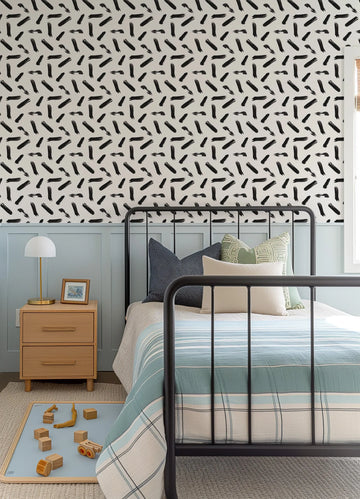 Lifestyle shot of a child's bedroom featuring "Gotta Dash" wallpaper with a repeating pattern of hand-painted black brushstrokes on a white background. The wallpaper covers the upper half of the wall, above light blue wainscoting. A black metal bed frame with a striped blue and white duvet and various pillows is centered in the shot. A wooden nightstand with a white lamp and framed photo sits to the left, and a blue playmat with wooden toys is on the floor.