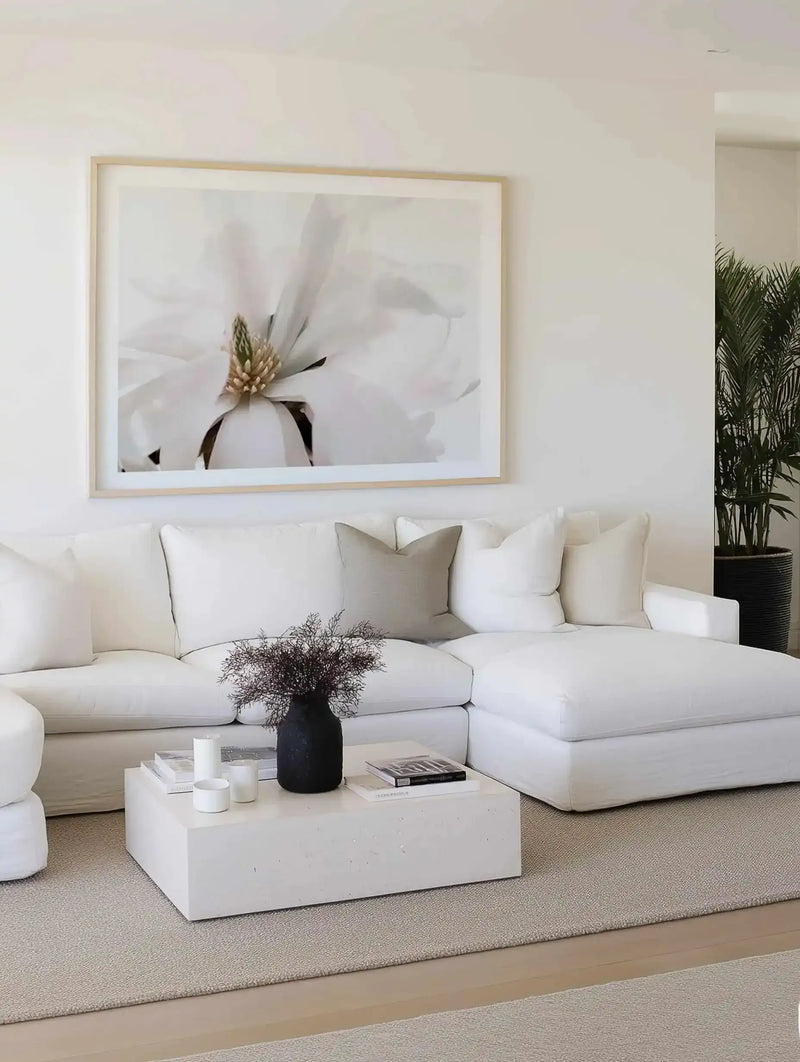 Lifestyle shot of a minimalist living room featuring a large framed art print of a white magnolia flower above a white sectional sofa. The art print is a close-up photograph of a white magnolia with a prominent green and beige center, displayed in a natural wood frame with a white border. The sofa has a light grey throw pillow and a black vase with dried dark foliage on a white coffee table. A tall green plant in a black pot is visible in the background.