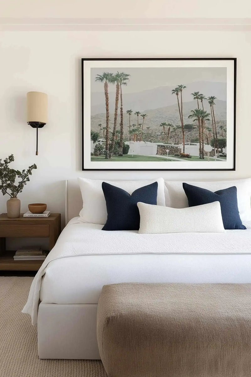 Framed art print displayed above a white upholstered bed in a modern bedroom. The photograph shows a Palm Springs landscape with tall palm trees, a white modernist building, manicured green lawn, and hazy mountains in the background under overcast sky. Black frame with white border. Bedroom features white bedding, dark navy and cream pillows, wood nightstand with ceramic vase and small plant, wall-mounted sconce with cream cylinder shade, and neutral textured area rug.
