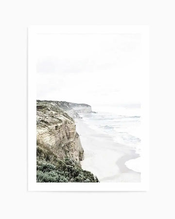Framed photograph of the Great Ocean Road coastline with a white frame and white border. The composition features a long stretch of sandy beach with gentle waves breaking, bordered by rugged, light-colored cliffs covered in sparse green vegetation under an overcast sky.