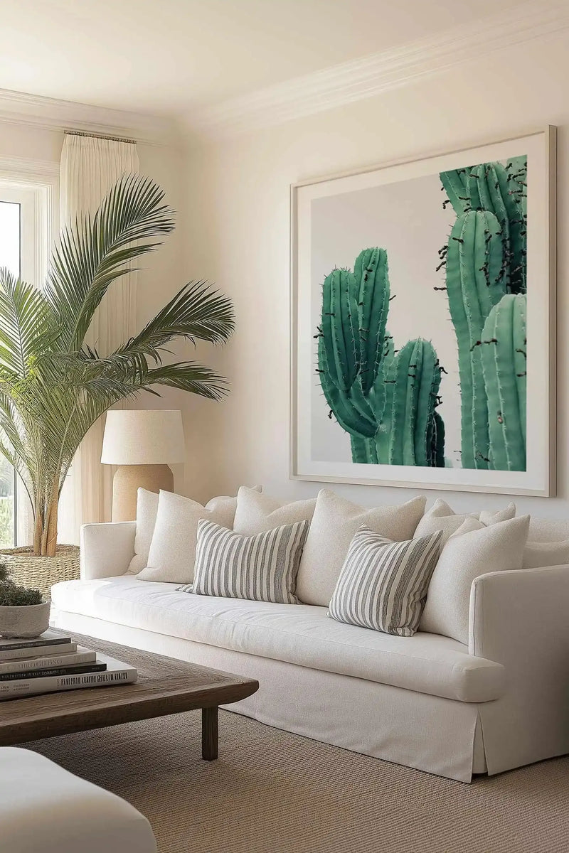 Framed art print displaying a close-up photograph of tall green columnar cacti against a soft beige background. The print features natural, realistic photography with rich sage and forest green tones showing the cacti's textured surface and small dark spines. The artwork is mounted in a light gray-toned frame with a white border and displayed on a cream-colored wall above a cream linen sofa styled with striped and solid neutral throw pillows in a bright, minimalist living room.