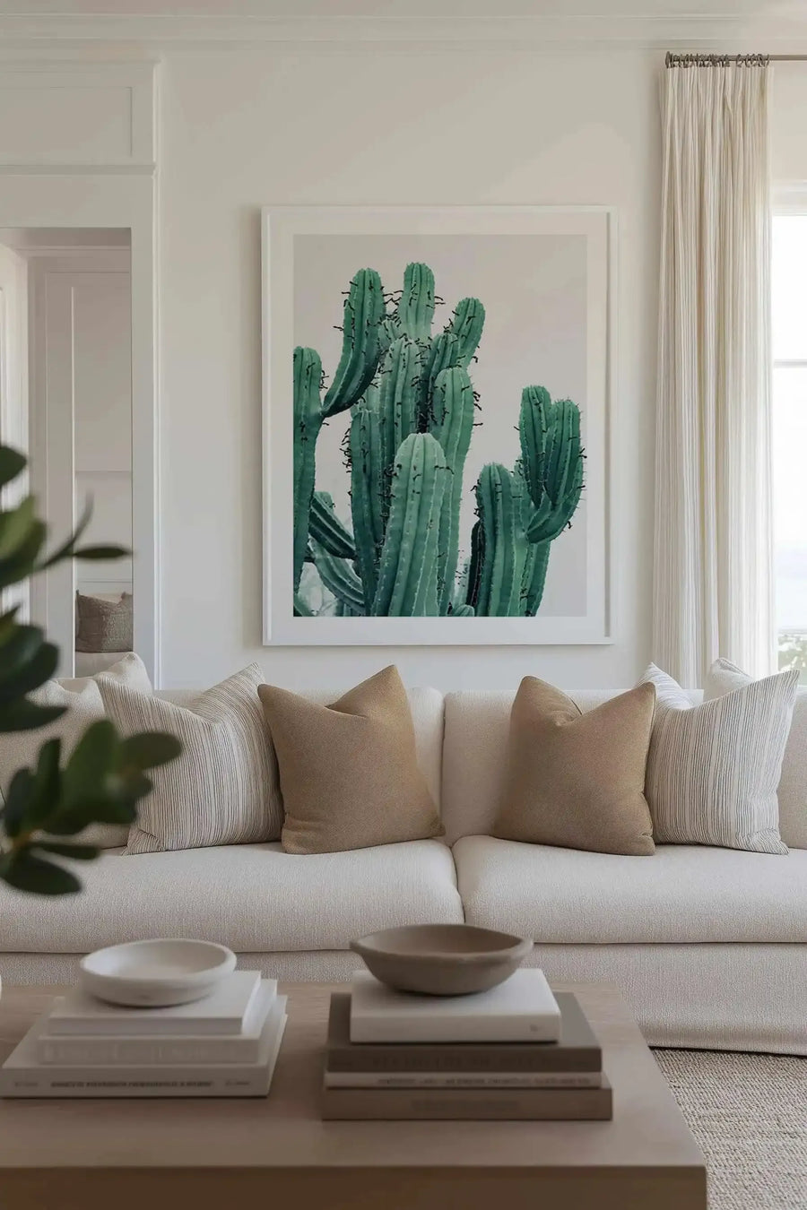 Lifestyle shot of a minimalist living room featuring a large framed art print of a cactus. The print depicts several tall, columnar cacti in varying shades of deep teal and emerald green, with visible spines, against a light cream background. The art print is displayed above a cream-coloured sofa adorned with striped and solid tan throw pillows. A light wood coffee table in the foreground holds decorative books and ceramic bowls.