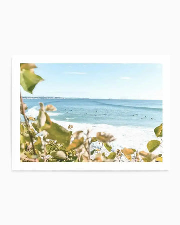 Photography art print of a coastal beach scene with a clear blue sky and gentle waves. The composition features numerous surfers scattered across the water, with lush green foliage in the foreground creating a natural frame. The distant coastline is visible on the left, and the overall mood is bright and serene.