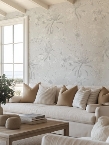 Lifestyle shot of a living room featuring Grey Banksia peel and stick wallpaper. The wallpaper features a repeating pattern of hand-painted Australian native Banksia flowers and foliage in muted grey tones on a light grey background, with a soft watercolour quality. The wallpaper is displayed on a wall with a large window looking out to a beach and ocean. A cream sofa with tan and striped cushions is in the foreground.