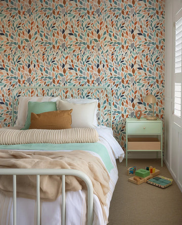 "Groovy Leaves wallpaper featuring a repeating pattern of scattered, overlapping leaves in various shades of teal, sage green, burnt orange, terracotta, peach, and grey on a cream background, displayed in a child's bedroom. The wallpaper covers the wall behind a mint green metal bed frame with white and striped bedding, a mint green throw, and a brown pillow. A mint green bedside table with a wooden drawer and a mushroom-shaped lamp sits to the right, and wooden toys are scattered on the carpeted floor."