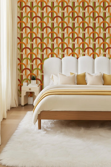 Lifestyle shot of a bedroom featuring Groovy Arches Wallpaper behind a bed with a white upholstered headboard. The wallpaper features a repeating pattern of semi-circles and vertical stripes in a retro-inspired palette of burnt orange, olive green, and warm beige on a cream background. The bed is dressed in cream bedding with a mustard yellow throw and pillows. A white fluffy rug covers the light wood floor, and a light beige nightstand with a gold knob holds a potted plant and books.