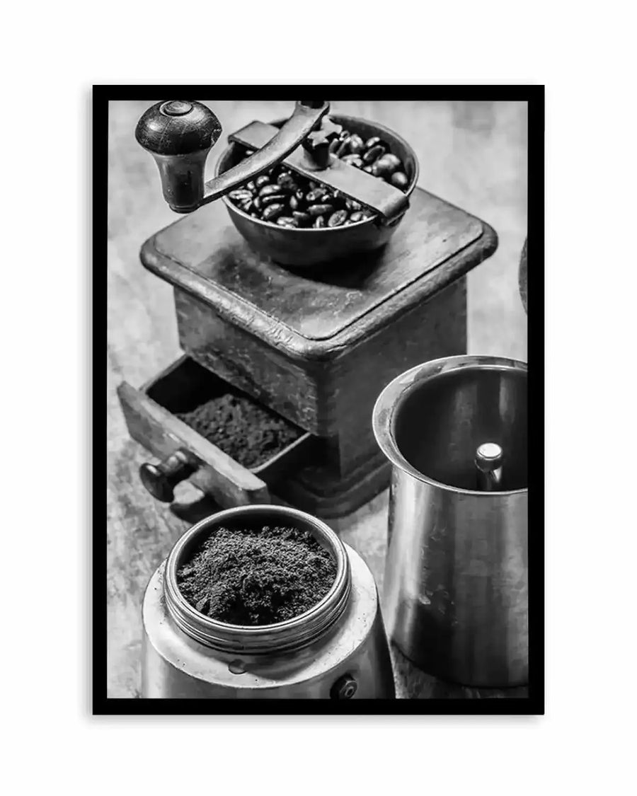Black and white overhead photograph of vintage coffee-making equipment and ground coffee, displayed as a framed art print with black border. The composition features a manual coffee grinder at top center filled with whole beans, a drawer of ground coffee below it, a glass jar containing finely ground coffee in the foreground, and a metal pitcher to the right. The monochromatic photography captures fine textures and details of the vintage metal and ceramic surfaces, creating a nostalgic, artisanal aesthetic.