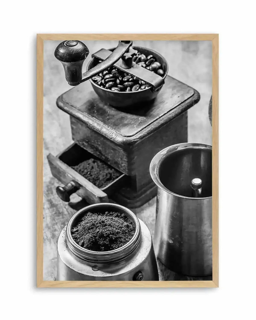 Black and white vintage photograph showing coffee preparation tools arranged vertically. At top, a manual coffee grinder with whole beans visible in its hopper and ground coffee in the catch drawer below. In the center, a metal measuring scoop and an open metal canister. At bottom, a glass jar filled with finely ground coffee and a dark metal pitcher. The composition showcases the texture and details of each implement against a neutral background. Displayed in a natural wood frame with white border.