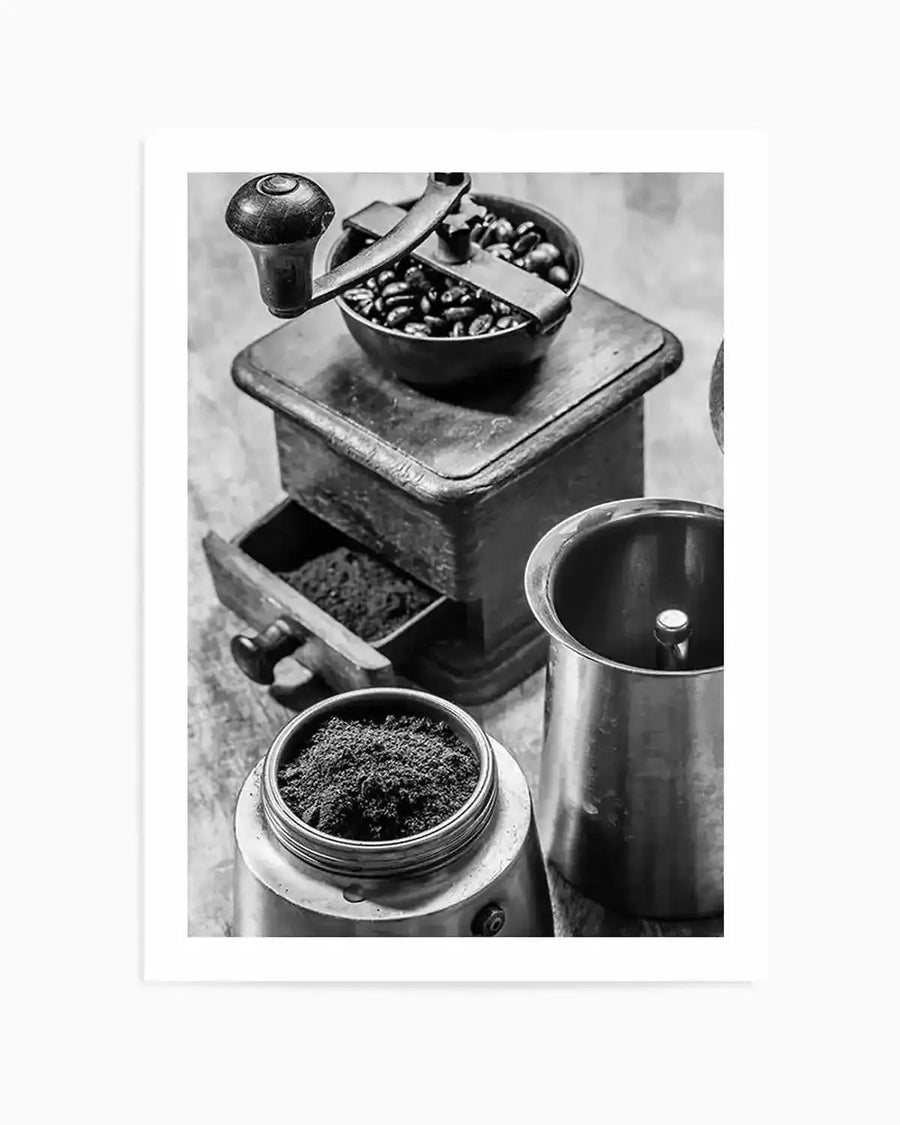 Framed black and white photograph of coffee preparation essentials. The composition features an antique coffee grinder with coffee beans in its hopper and ground coffee in its drawer, alongside a disassembled Moka pot with ground coffee in its filter basket and the lower chamber visible.