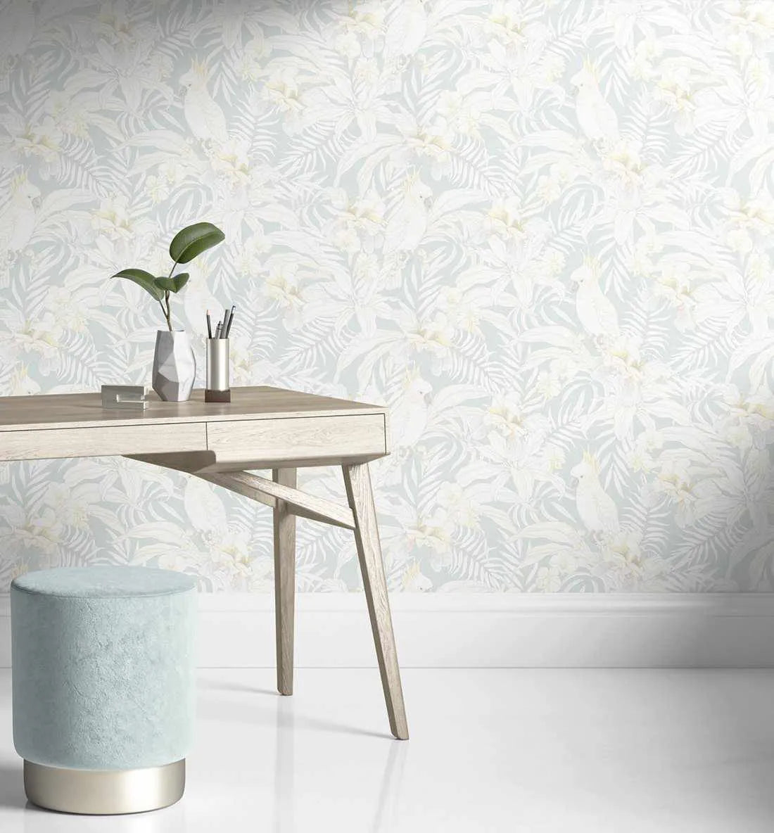 Home Office:
"Peel and stick wallpaper featuring a repeating pattern of white cockatoos, large tropical leaves, and orchids in soft cream and pale yellow, set against a light blue background. The wallpaper is displayed in a home office with a light wood desk, a grey geometric vase with a green plant, a silver pencil holder, and a light blue velvet stool with a gold base."