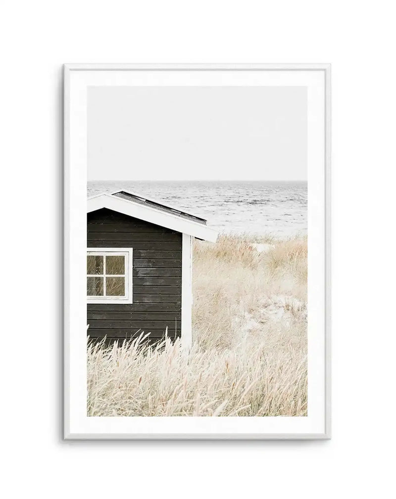 Framed black and white photograph of a small, dark wooden beach hut with a white-trimmed roof and window, set amidst tall, light-coloured dune grasses. The ocean with gentle waves is visible in the background under a bright, overcast sky. The print is displayed in a white frame with a white border.