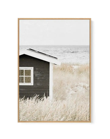 Framed canvas art print featuring a coastal scene with a dark charcoal wooden beach hut with white-trimmed window positioned in dried beige beach grass. Behind the hut, calm grey-blue ocean waters meet a pale overcast sky at the horizon. A white boat is partially visible in the upper right. Natural oak wood frame with white border surrounding the photograph-style image.