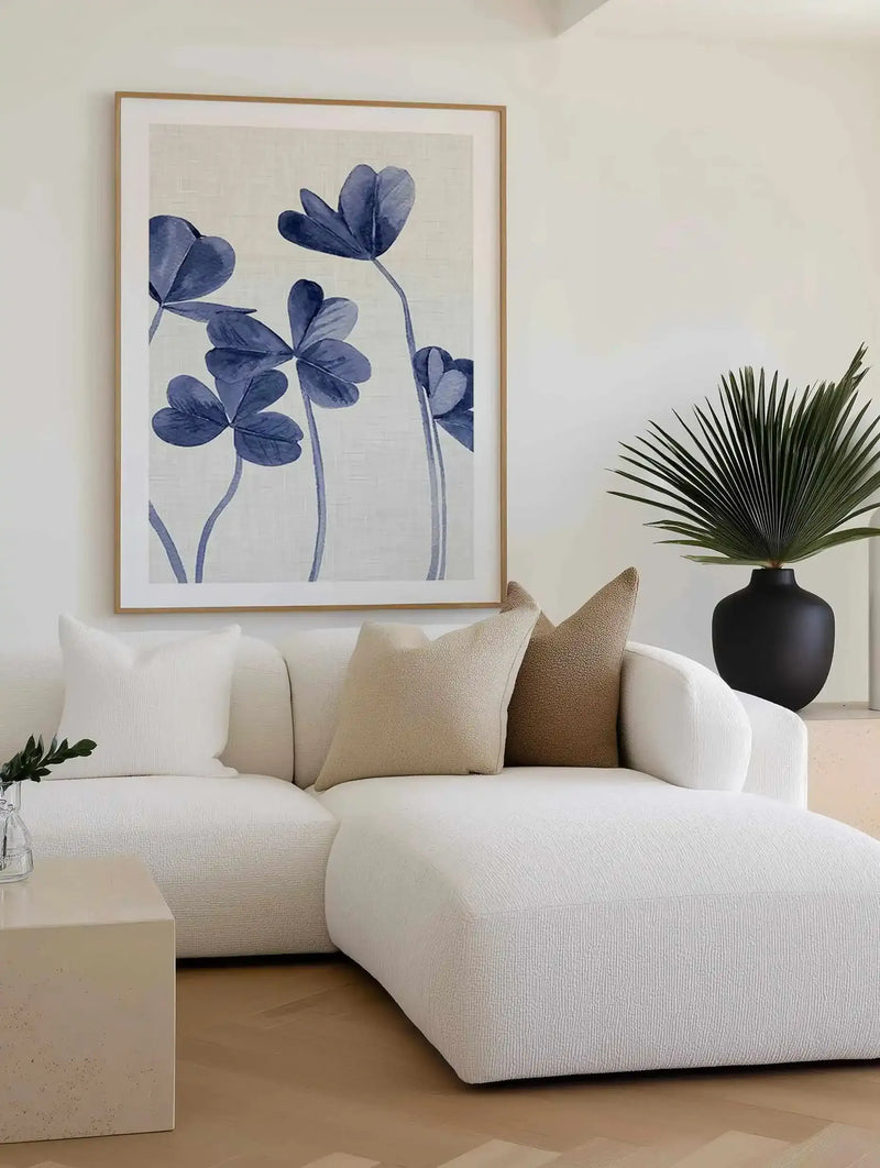 Lifestyle shot of a minimalist living room featuring a large framed art print displayed above a curved white sofa. The print features five stylized clover-like leaves in varying shades of indigo and periwinkle blue with slender stems, on a textured cream background, with a natural wood frame and white border. The sofa is adorned with cream and beige textured throw pillows, and a black vase with a large green palm frond sits on a light-colored side table.