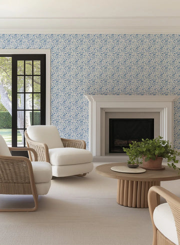 "Hamptons Hydrangea wallpaper featuring a repeating pattern of small, hand-painted hydrangea florets in varying shades of blue, from light sky blue to deep navy, on a soft white background. The wallpaper is displayed in a bright living room above a white fireplace mantel and next to a black-framed window overlooking a garden. Two cream-coloured armchairs with natural wicker frames and a round wooden coffee table with a potted plant are visible in the foreground."