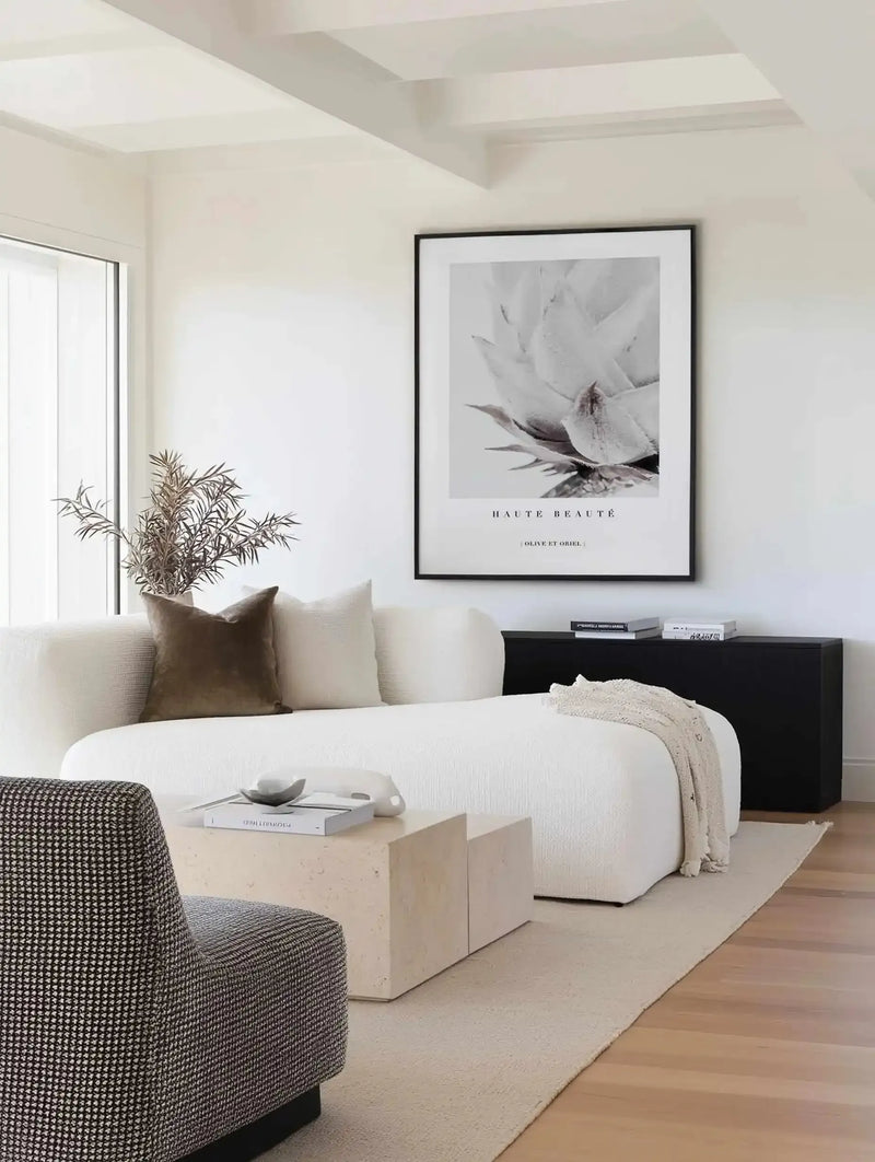 Framed black and white photographic art print displayed above a black nightstand in a minimalist bedroom. The print features a close-up of a King Protea flower with delicate, layered petals and visible stamens in grayscale tones. Black frame with white border. Text reads "HAUTE BEAUTE" and "OLIVE ET ORIEL" in serif font below the image. Room features white walls, cream upholstered furniture, natural wood flooring, and a potted plant by the window.