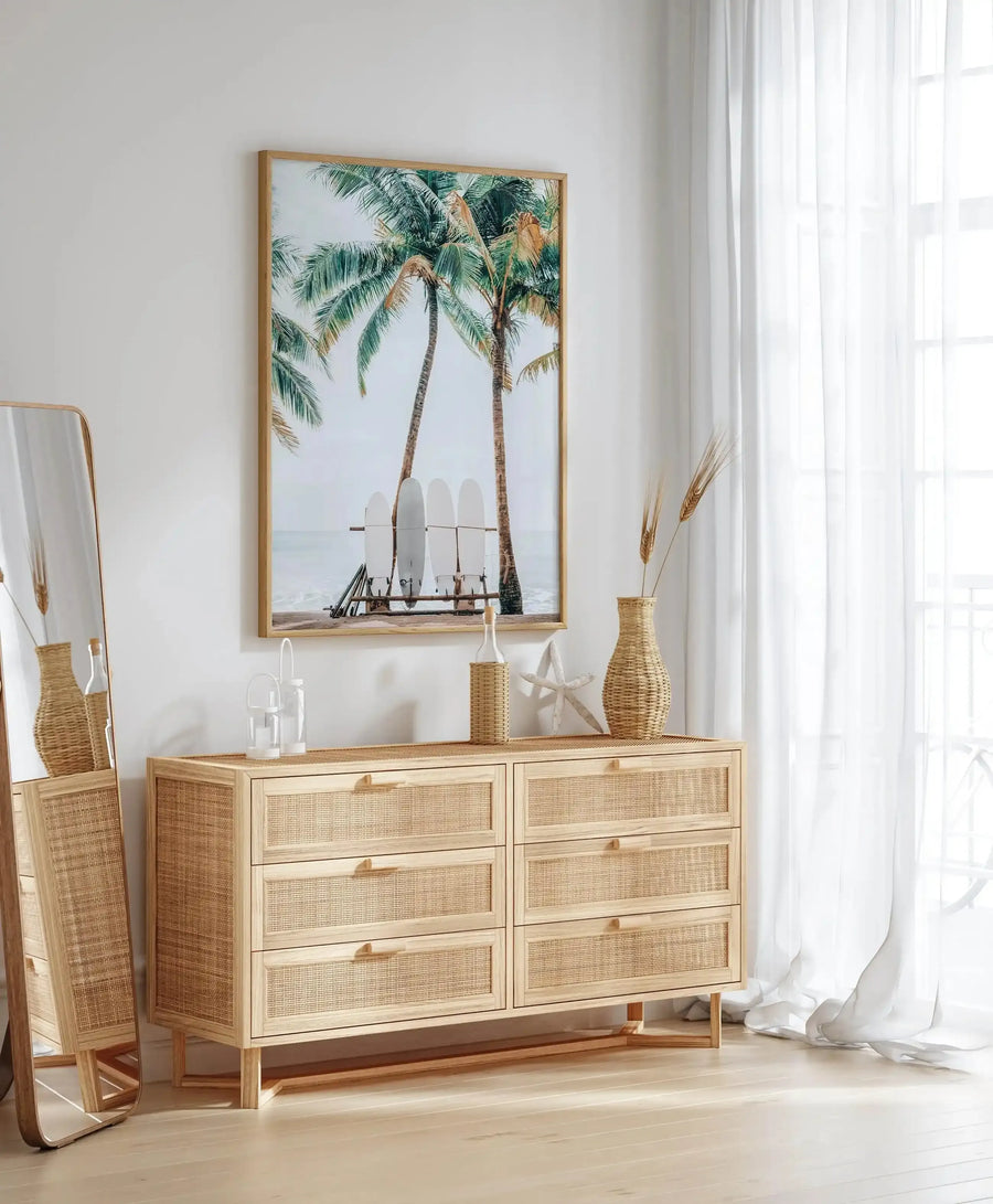 Lifestyle shot of a bedroom featuring a framed art print displayed above a light wood and rattan dresser. The print is a photograph of a tropical beach scene with two tall palm trees and four white surfboards leaning against a wooden rack on the sand, with the ocean in the background. The print has a natural wood frame. The room includes a full-length mirror with a wood frame, a woven vase with dried wheat stalks, a clear bottle with a woven sleeve, a white starfish, and white sheer curtains.