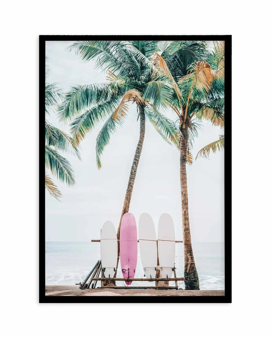 Framed art print featuring a photograph of four surfboards leaning on a wooden rack beneath two tall palm trees on a sandy beach. One surfboard is dusty rose pink while three are white. The ocean and pale overcast sky are visible in the background. The photograph has a soft, muted colour palette with natural textures. Black frame with white border.