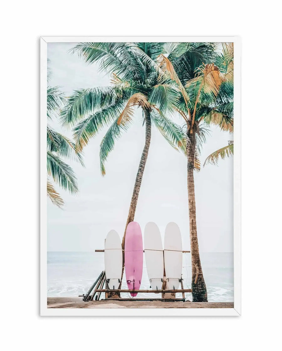 Framed art print featuring a photograph of surfboards leaning against a wooden rack between two tall palm trees on a sandy beach. The composition includes four surfboards—three in white and one in dusty rose pink—positioned vertically beneath swaying palm fronds with green and golden-brown foliage. The ocean and light overcast sky are visible in the soft-focus background. The image has a calm, tropical aesthetic with natural, muted tones. White wooden frame with white border.