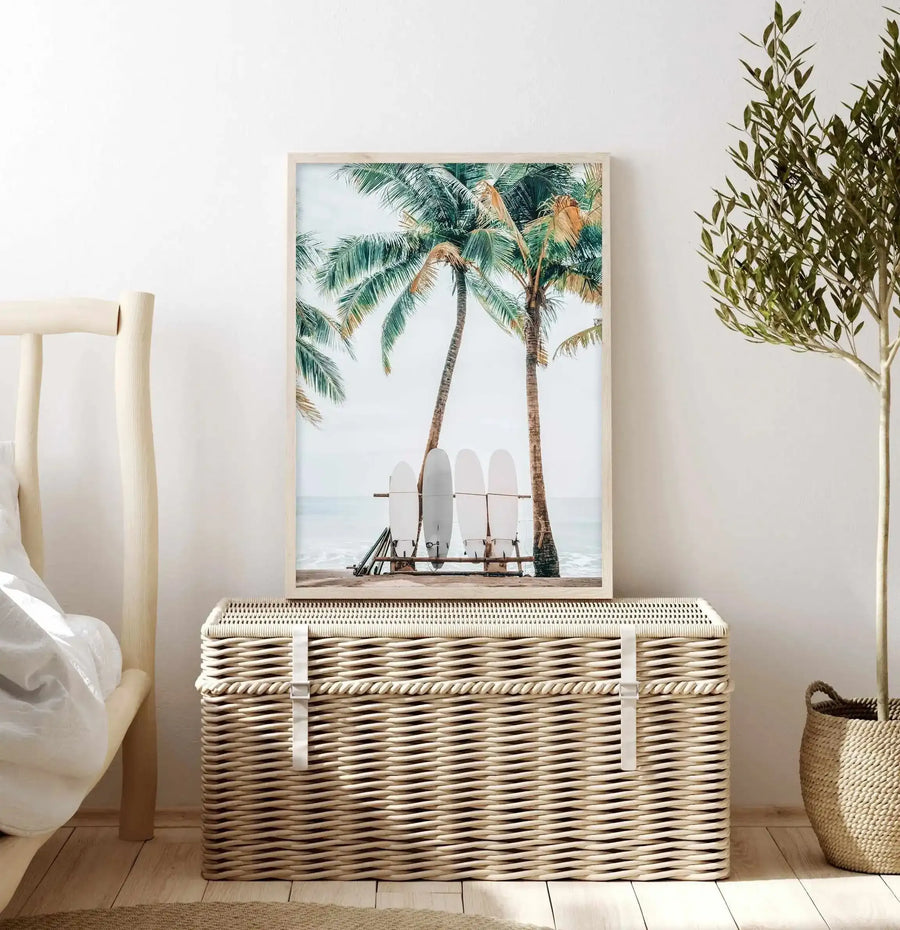Lifestyle shot of a bedroom with a framed art print displayed on a woven trunk. The print features a tropical beach scene with four white surfboards leaning against two tall palm trees with green and golden fronds, with the ocean and a light sky in the background. The room includes a light wood bed frame with white bedding on the left, a large woven trunk in the center, and a tall plant with slender green leaves in a woven basket on the right.