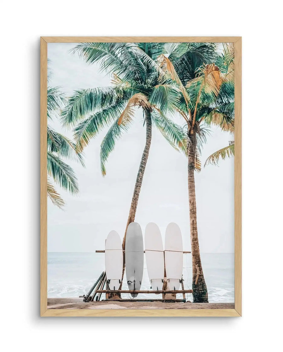 Framed art print featuring a coastal scene with four white surfboards leaning against a wooden rack between two tall palm trees on a sandy beach. The ocean and overcast sky are visible in the background. Palm fronds with green and golden-brown tones frame the top of the composition. The artwork has a soft, muted color palette of whites, greens, tans, and grays with a photographic style. Natural wood frame with white border.