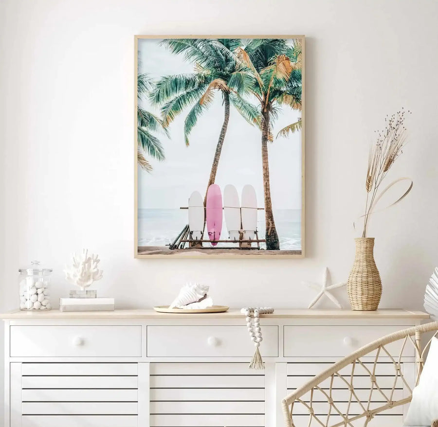 Framed art print featuring a coastal scene with tall palm trees, surfboards leaning against a wooden rack, and calm ocean waters beneath a light sky. The composition centers a pink surfboard among white and neutral-toned boards. A natural wood frame with white border displays the photograph-style artwork in a minimalist coastal-themed bedroom setting, positioned above a white dresser decorated with woven vase, dried grasses, shells, and decorative objects.