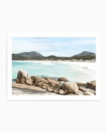 Framed photograph of a serene coastal beach scene with a white border. The composition features a turquoise ocean with gentle white waves breaking on a white sand beach, backed by green dunes and distant grey mountains under a pale blue sky. Large, smooth, light brown boulders are visible in the foreground along the water's edge.