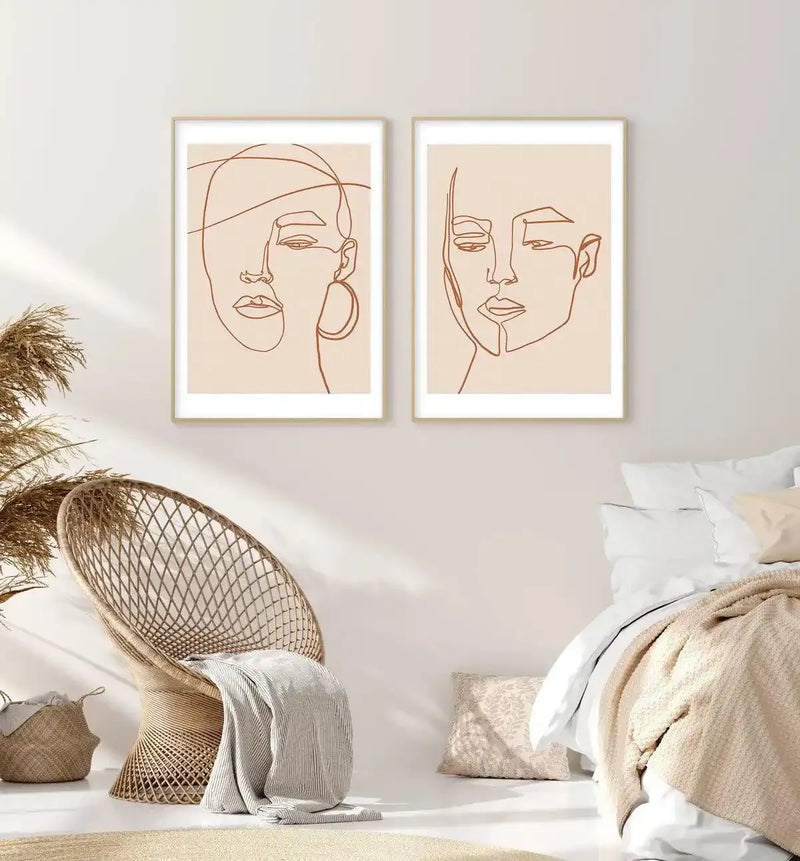 Lifestyle shot of a bedroom featuring two framed art prints above a bed and a woven chair. The prints feature continuous line art portraits of two women in terracotta brown on a cream background, displayed in natural wood frames with white borders. The room includes a beige woven chair with a striped cream throw, a woven basket with dried pampas grass, and a bed with white and beige bedding.