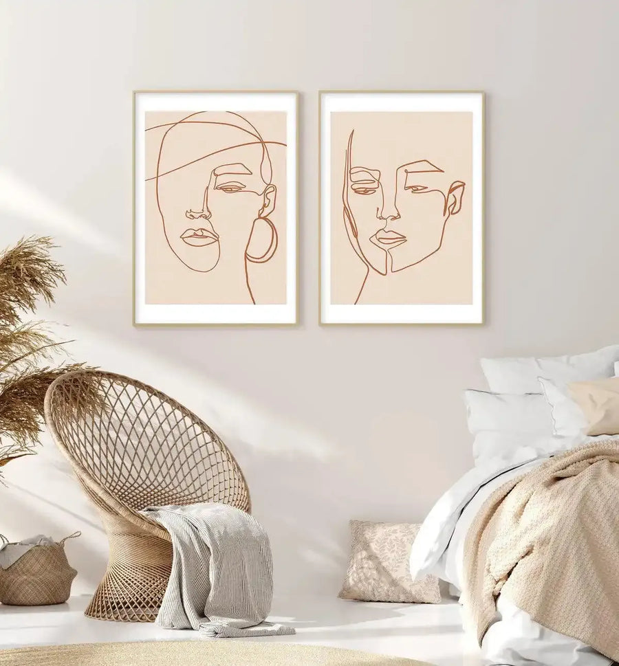 Lifestyle shot of a bedroom featuring two framed art prints above a bed and a woven chair. The prints feature continuous line art portraits of two women in terracotta brown on a cream background, displayed in natural wood frames with white borders. The room includes a beige woven chair with a striped cream throw, a woven basket with dried pampas grass, and a bed with white and beige bedding.