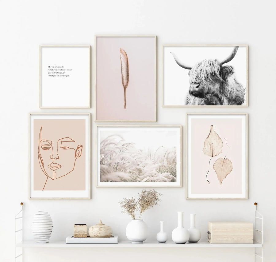 Lifestyle shot of a gallery wall featuring six framed art prints above a white shelf with decorative objects. The product, "Her Contours | Terracotta Art Print," is a framed line art illustration of a woman's face in terracotta orange on a light peach background, with a natural wood frame and white border. Other prints include a quote, a feather, a Highland cow, pampas grass, and dried leaves. The shelf holds white vases, a white tray with a lidded pot, and a wooden box.