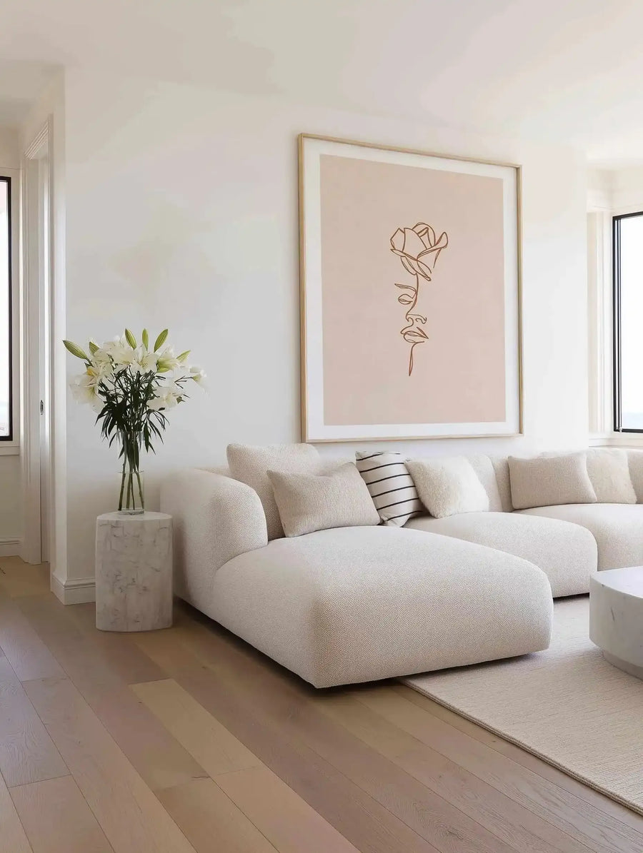 Large framed art print displayed above a curved white sofa in a minimalist living room. The print features a single line drawing of a rose and a woman's face in terracotta against a warm cream background, with a natural wood frame and white border. The room includes a textured cream throw, a striped cushion, and white lilies in a glass vase on a marble side table.
