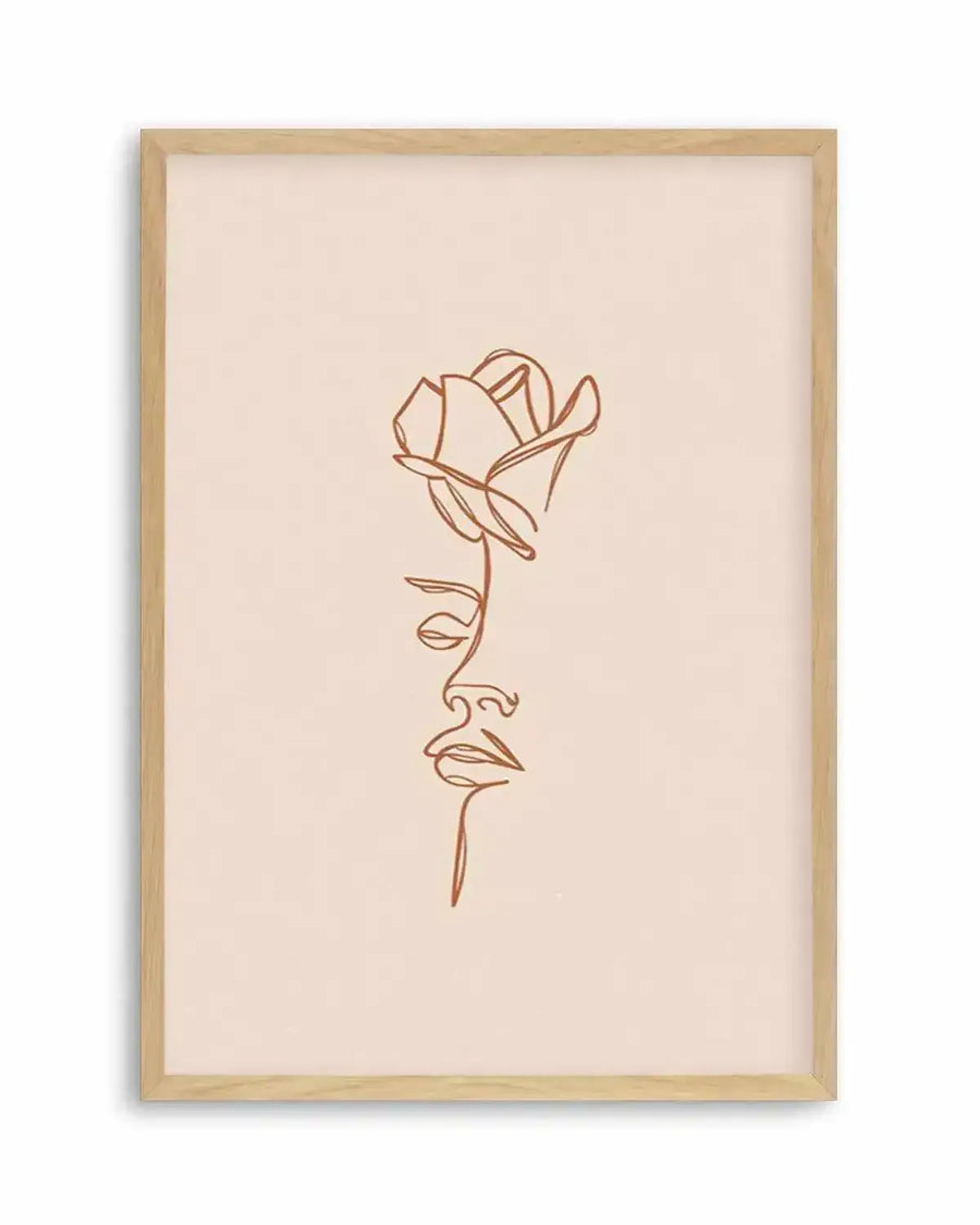 Line art print featuring a single continuous terracotta line forming the profile of a woman's face with a rose blooming from her head, on a pale peach background, displayed in a natural oak frame with white border.