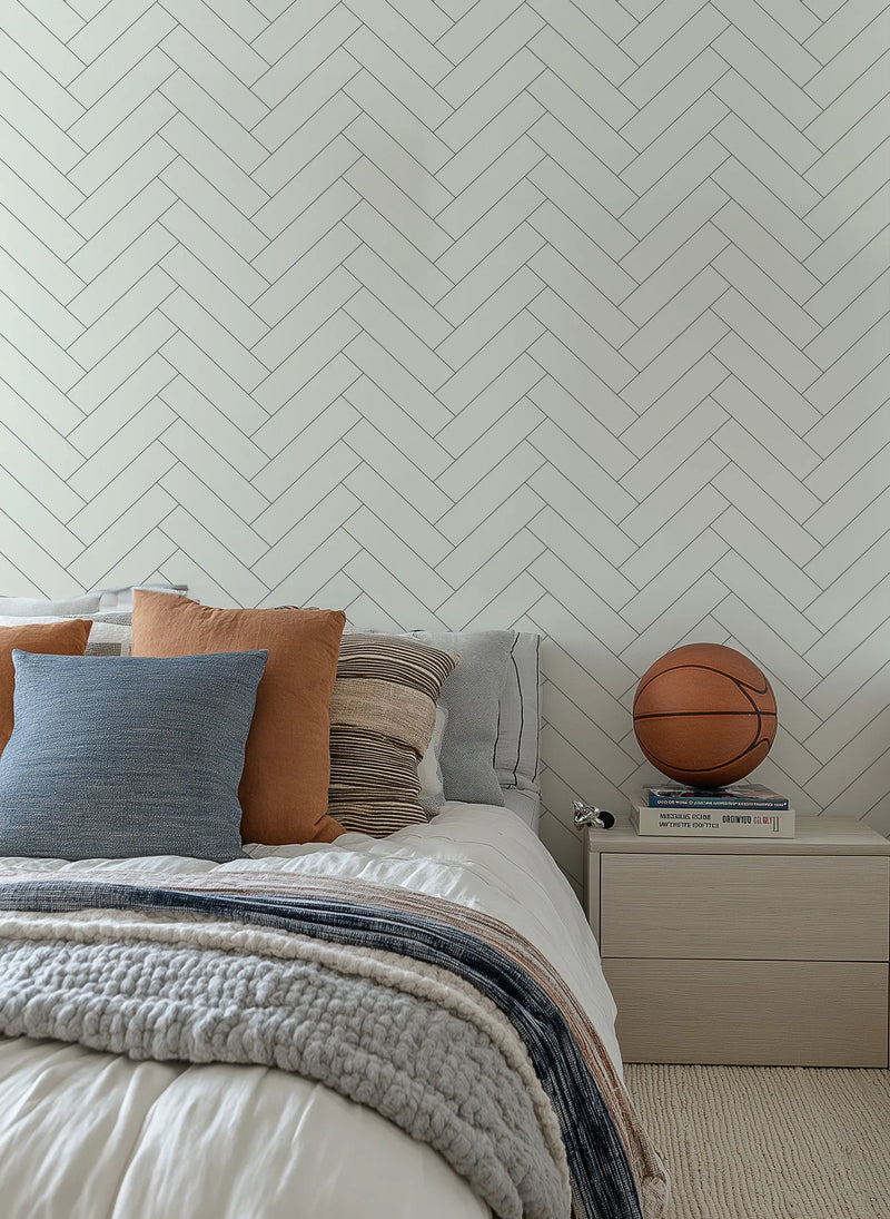 Herringbone in Stone wallpaper displayed on a bedroom wall behind a bed with decorative pillows and layered bedding. The wallpaper features a classic herringbone tile pattern in soft sage green and light gray tones with subtle linear detailing throughout each rectangular tile. The diagonal interlocking pattern creates visual movement across the wall. A basketball and stacked books sit on a light gray nightstand beside the bed.