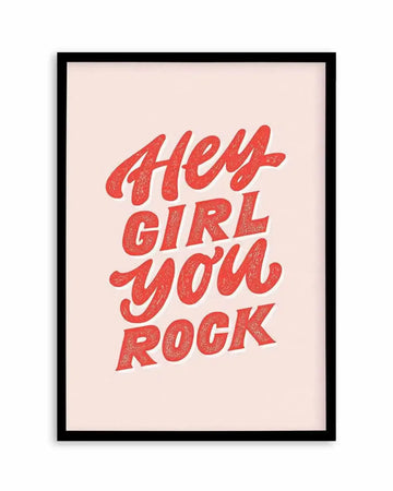 Art print featuring the phrase "Hey Girl You Rock" in a bold, hand-lettered script with a textured, distressed finish in a vibrant red hue, set against a soft, pale pink background, displayed in a black frame with a white border.
