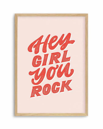 Art print featuring the phrase "Hey Girl You Rock" in bold, textured, hand-lettered red script with a subtle shadow effect, on a soft blush pink background, displayed in a natural oak frame with white border.