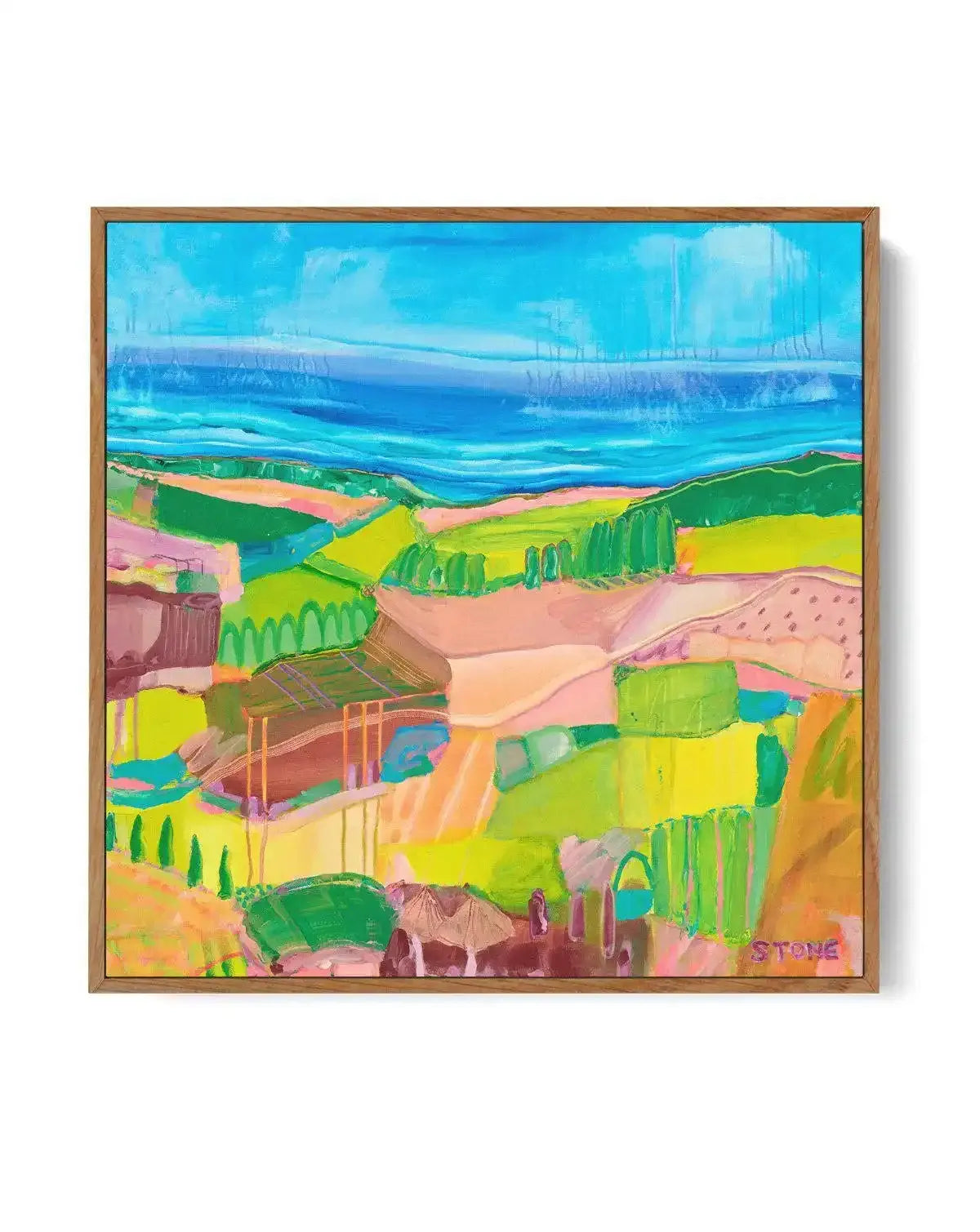 Abstract canvas print featuring a vibrant, expressive landscape with a bright blue sky and ocean in the background, and a foreground of rolling hills, fields, and structures in shades of lime green, yellow, pink, and brown, with visible brushstroke texture, displayed in a natural oak frame.